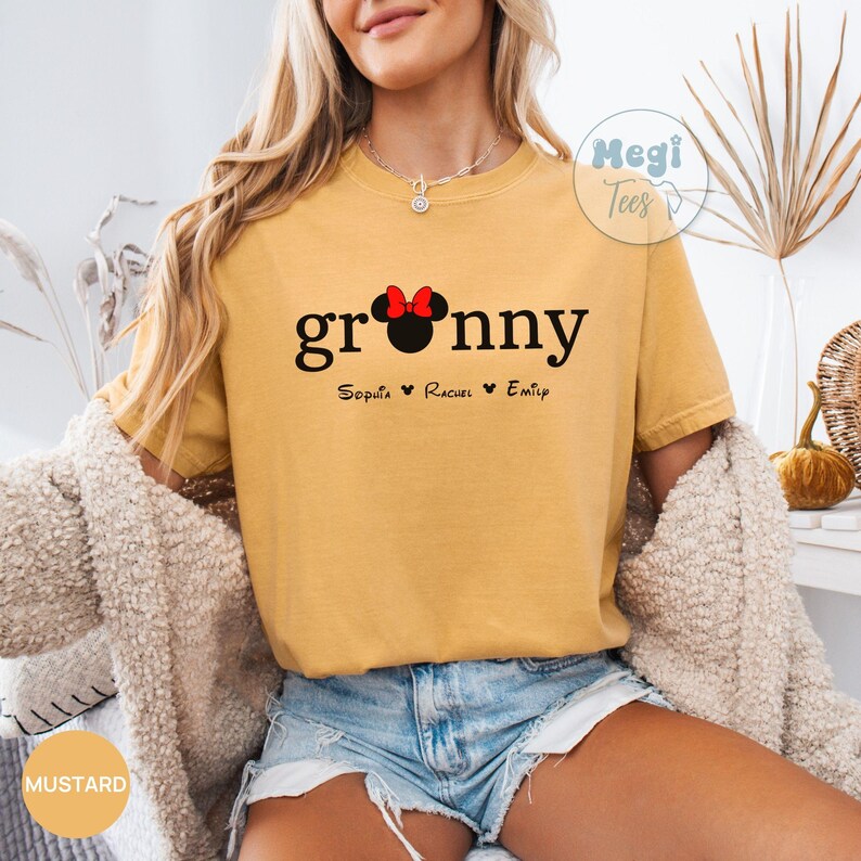 May include: Mustard yellow t-shirt with the word "granny" in black script, a Minnie Mouse head, and three names below. The shirt has a relaxed fit and is worn with denim shorts and a cream-colored cardigan.