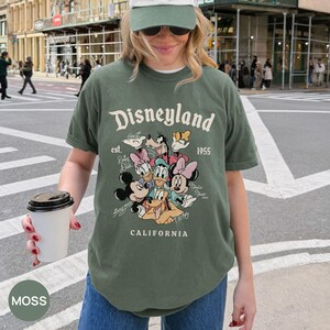 Puede incluir: Camiseta verde musgo con el logotipo de Disneyland y un gr&aacute;fico de personajes de Disney. La camiseta tiene el texto "Disneyland est. 1955 CALIFORNIA". Es una camiseta de manga corta con cuello redondo.