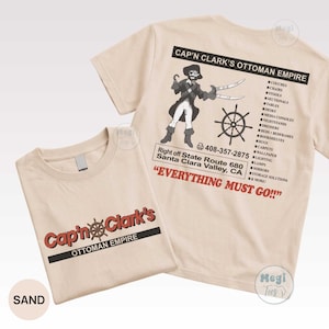 May include: A sand-colored t-shirt featuring a graphic design. The front displays the text Cap'n Clark's Ottoman Empire. The back shows a pirate illustration, a ship wheel, the text Cap'n Clark's Ottoman Empire, a list of furniture, and Everything Must Go.