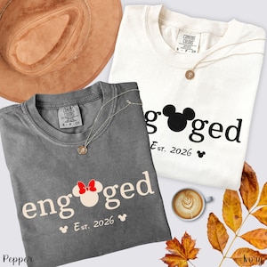 K&ouml;nnte beinhalten: Zwei T-Shirts, eines in Grau und eines in Creme, mit dem Wort "engaged" und einem Mickey-Mouse-Kopf-Design. Die Shirts zeigen auch "Est. 2026" und ein kleines Mickey-Mouse-Symbol. Ein brauner Hut, eine Halskette und eine Kaffeetasse sind ebenfalls im Bild.