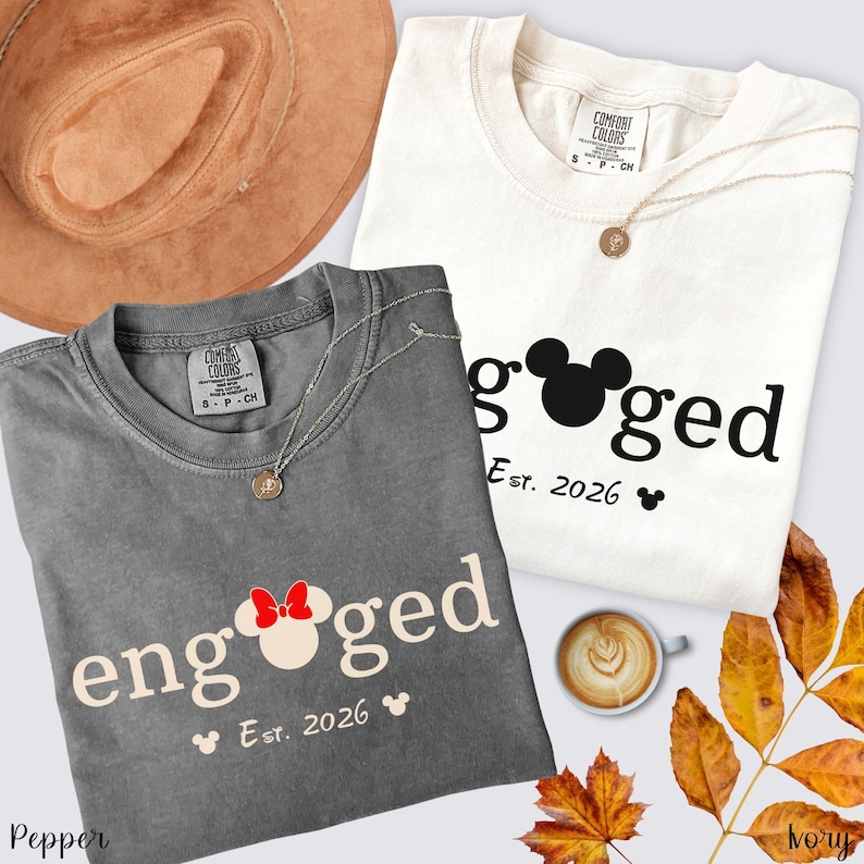K&ouml;nnte beinhalten: Zwei T-Shirts, eines in Grau und eines in Creme, mit dem Wort "engaged" und einem Mickey-Mouse-Kopf-Design. Die Shirts zeigen auch "Est. 2026" und ein kleines Mickey-Mouse-Symbol. Ein brauner Hut, eine Halskette und eine Kaffeetasse sind ebenfalls im Bild.
