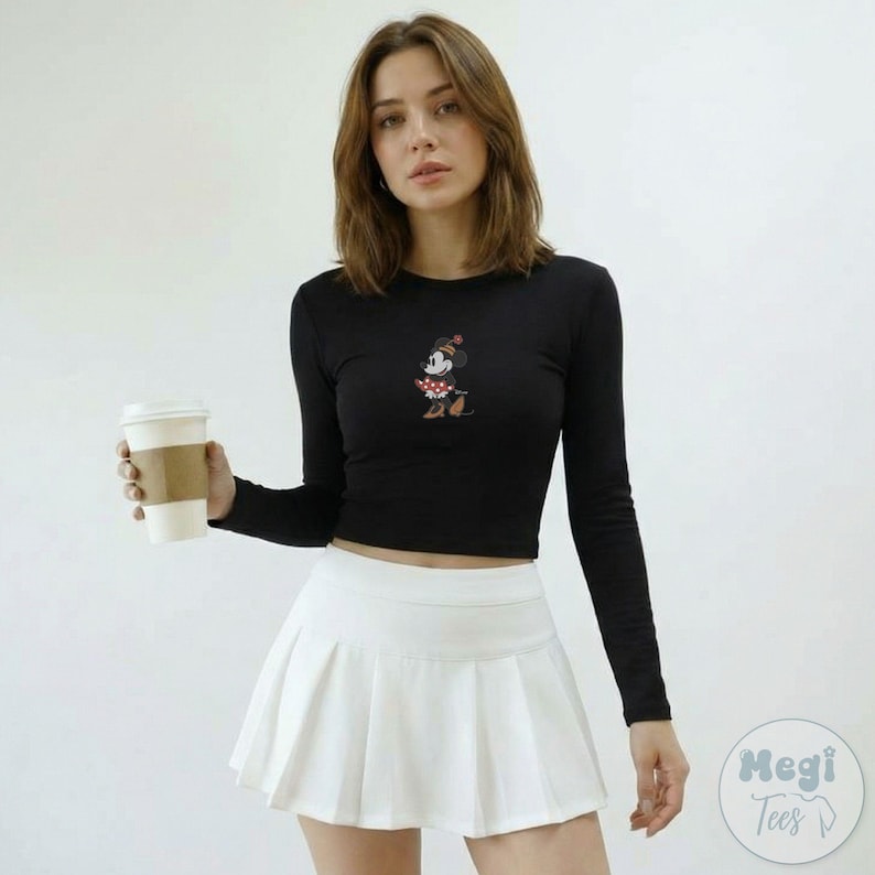 Puede incluir: Top corto negro de manga larga con un dise&ntilde;o de Minnie Mouse, combinado con una falda plisada blanca. El top tiene cuello redondo y el dise&ntilde;o de Minnie Mouse est&aacute; centrado. La falda es de estilo tenis cl&aacute;sico.