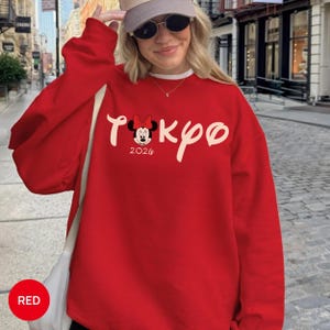 May include: A red sweatshirt with the word "TOKYO" in white script and a Minnie Mouse graphic. The year "2026" is printed below the graphic. The sweatshirt has long sleeves and a crew neck.