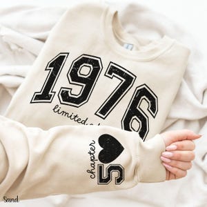 May include: Sand-colored sweatshirt with the year "1976" printed in large, black, collegiate-style lettering. The sleeve features the text "chapter 55" with a black heart. The sweatshirt is a casual, comfortable style.