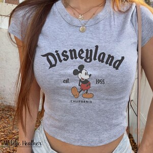 May include: Gray crop top with the Disneyland logo, Mickey Mouse graphic, and the text "est. 1955 CALIFORNIA". The shirt is made of Athletic Heather material.