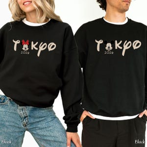 May include: Black sweatshirts with white crew necks. The sweatshirts feature the word "Tokyo" in a stylized font, with a Mickey Mouse graphic and the year "2026". One sweatshirt has a Minnie Mouse graphic with a red bow.