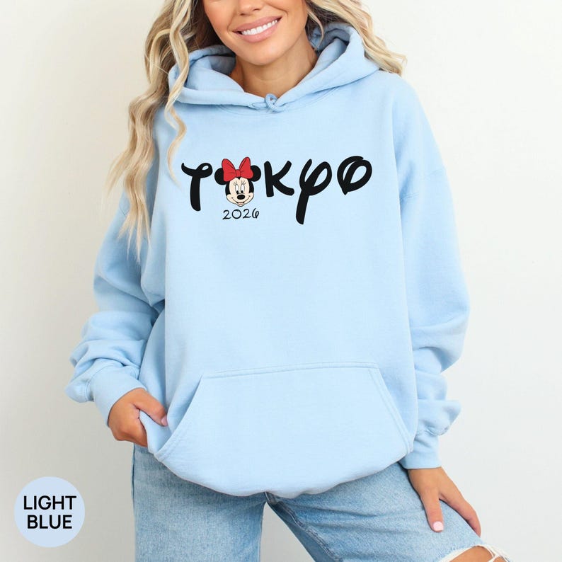 May include: Light blue hoodie with the word "Tokyo" in black script, featuring a Minnie Mouse graphic with a red bow. The year "2026" is below the graphic. The bottom left corner of the hoodie has the words "LIGHT BLUE".