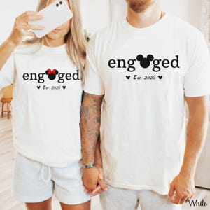 K&ouml;nnte beinhalten: Zwei wei&szlig;e T-Shirts mit dem Wort "engaged" in Schwarz aufgedruckt. Ein Shirt hat eine rote Minnie-Maus-Schleife, das andere einen Mickey-Mouse-Kopf. Beide Shirts haben "Est. 2026" darunter aufgedruckt. Die Shirts werden von einem Paar getragen.