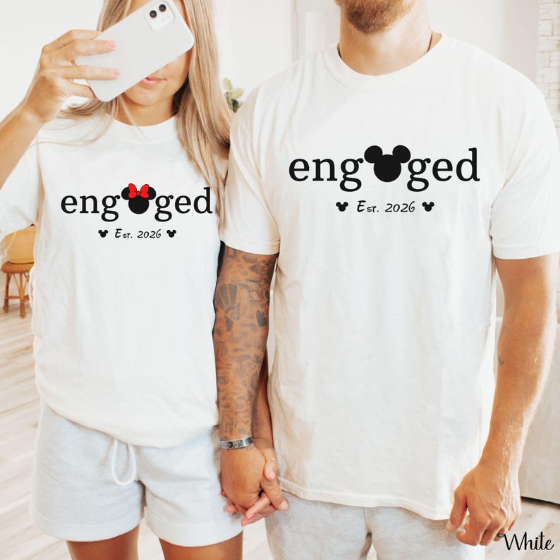 K&ouml;nnte beinhalten: Zwei wei&szlig;e T-Shirts mit dem Wort "engaged" in Schwarz aufgedruckt. Ein Shirt hat eine rote Minnie-Maus-Schleife, das andere einen Mickey-Mouse-Kopf. Beide Shirts haben "Est. 2026" darunter aufgedruckt. Die Shirts werden von einem Paar getragen.
