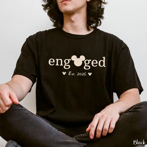 K&ouml;nnte beinhalten: Schwarzes T-Shirt mit dem Wort "engaged" in wei&szlig;er Schrift, mit einem Mickey Mouse-Kopf anstelle des "o". Darunter steht "Est. 2026" mit zwei kleinen Mickey Mouse-K&ouml;pfen. Das Shirt ist ein l&auml;ssiger Stil.