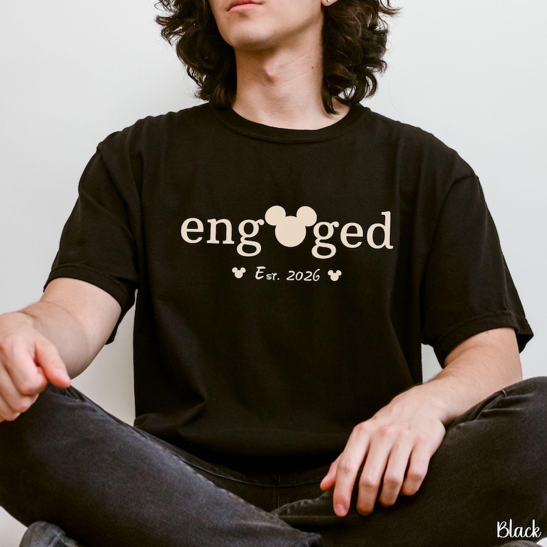 K&ouml;nnte beinhalten: Schwarzes T-Shirt mit dem Wort "engaged" in wei&szlig;er Schrift, mit einem Mickey Mouse-Kopf anstelle des "o". Darunter steht "Est. 2026" mit zwei kleinen Mickey Mouse-K&ouml;pfen. Das Shirt ist ein l&auml;ssiger Stil.