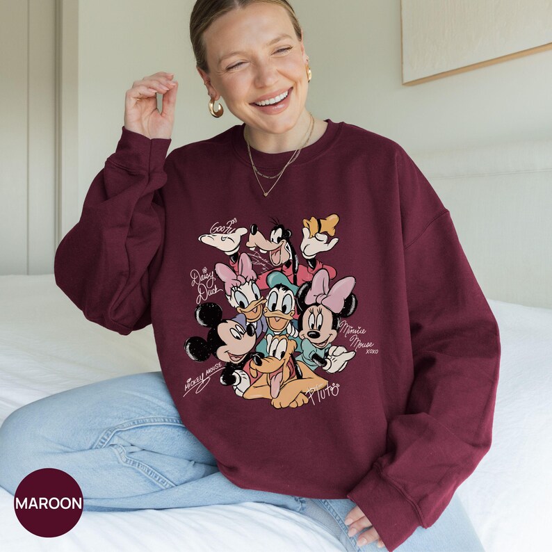 K&ouml;nnte beinhalten: Burgunderfarbenes Sweatshirt mit einer Grafik von Micky Maus, Minnie Maus, Donald Duck, Daisy Duck, Goofy und Pluto. Das Design enth&auml;lt die Namen der Charaktere in einer Schriftart. Das Sweatshirt hat eine einfarbige burgunderrote Farbe.