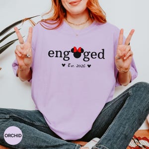 K&ouml;nnte beinhalten: Ein helllila T-Shirt mit dem Wort "engaged" in schwarzer Schrift. Das "o" in "engaged" wird durch einen Minnie Mouse-Kopf mit roter Schleife ersetzt. Darunter steht "Est. 2026" mit zwei kleinen schwarzen Herzen.