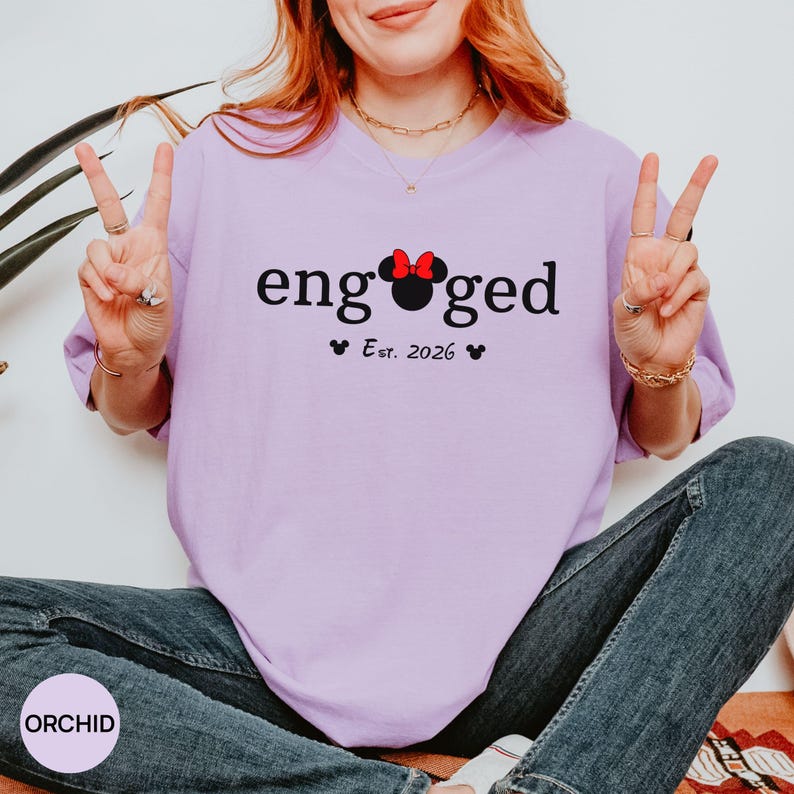 K&ouml;nnte beinhalten: Ein helllila T-Shirt mit dem Wort "engaged" in schwarzer Schrift. Das "o" in "engaged" wird durch einen Minnie Mouse-Kopf mit roter Schleife ersetzt. Darunter steht "Est. 2026" mit zwei kleinen schwarzen Herzen.