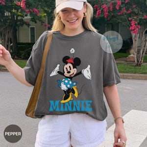 May include: Dark gray t-shirt featuring a graphic of Minnie Mouse in a playful pose, with a red bow, blue skirt, and yellow shoes. The word "MINNIE" is printed in blue below the character. The shirt is worn with white shorts.