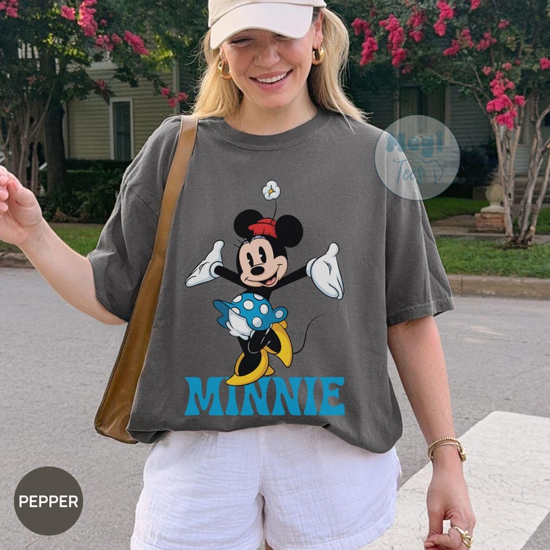 May include: Dark gray t-shirt featuring a graphic of Minnie Mouse in a playful pose, with a red bow, blue skirt, and yellow shoes. The word "MINNIE" is printed in blue below the character. The shirt is worn with white shorts.