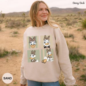 May include: A beige sweatshirt featuring four panels with Daisy Duck illustrations. Each Daisy Duck image has a leopard print bow. The sweatshirt has the text "Daisy Duck" in the upper right corner and "SAND" in the lower left corner.