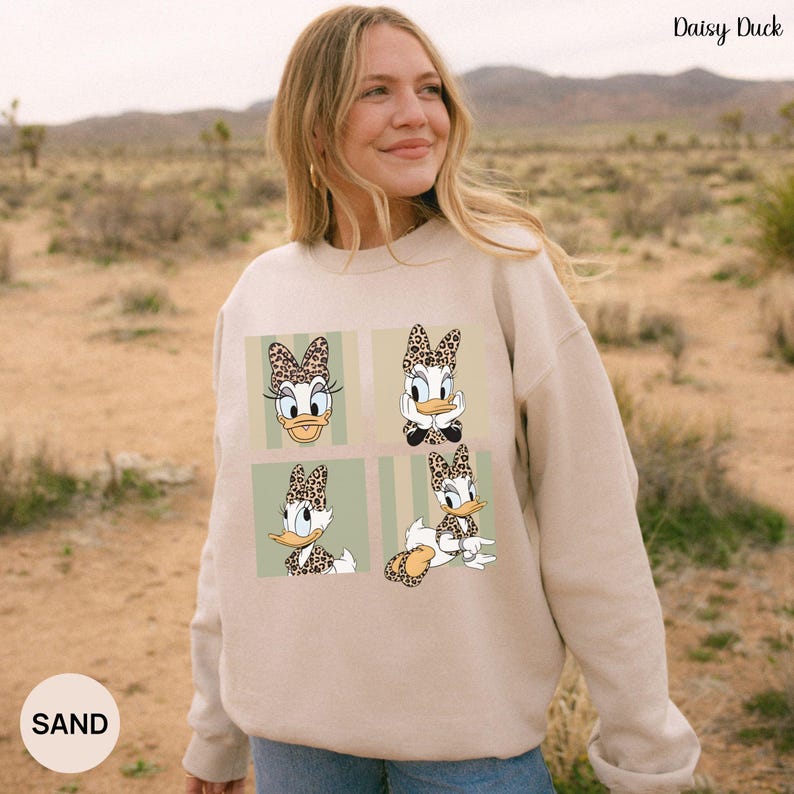 May include: A beige sweatshirt featuring four panels with Daisy Duck illustrations. Each Daisy Duck image has a leopard print bow. The sweatshirt has the text "Daisy Duck" in the upper right corner and "SAND" in the lower left corner.