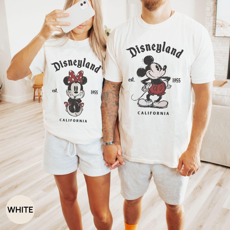 Puede incluir: Dos camisetas blancas con el logotipo de Disneyland y gr&aacute;ficos de Mickey y Minnie Mouse. Las camisetas tienen el texto "est. 1955 CALIFORNIA". La persona de la izquierda lleva pantalones cortos grises, y la persona de la derecha lleva pantalones cortos grises y calcetines naranjas.