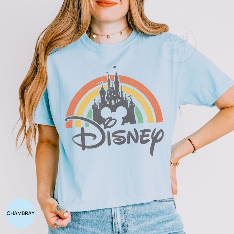 May include: Light blue t-shirt featuring a colorful rainbow design with a castle silhouette, Mickey Mouse ears, and the word "Disney" in a stylized font. The shirt is made of a soft material and is a comfortable fit.