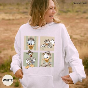 May include: White hooded sweatshirt featuring a four-panel graphic of Donald Duck in safari attire. The top right corner of the sweatshirt has the text "Donald Duck". The bottom left corner of the sweatshirt has the text "WHITE".
