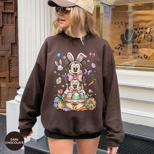 Puede incluir: Sudadera marr&oacute;n chocolate oscuro con un gr&aacute;fico colorido de Pascua. El gr&aacute;fico incluye los personajes de Mickey y Minnie Mouse con orejas de conejo, huevos de Pascua, zanahorias y mariposas. La sudadera tiene cuello redondo y mangas largas.