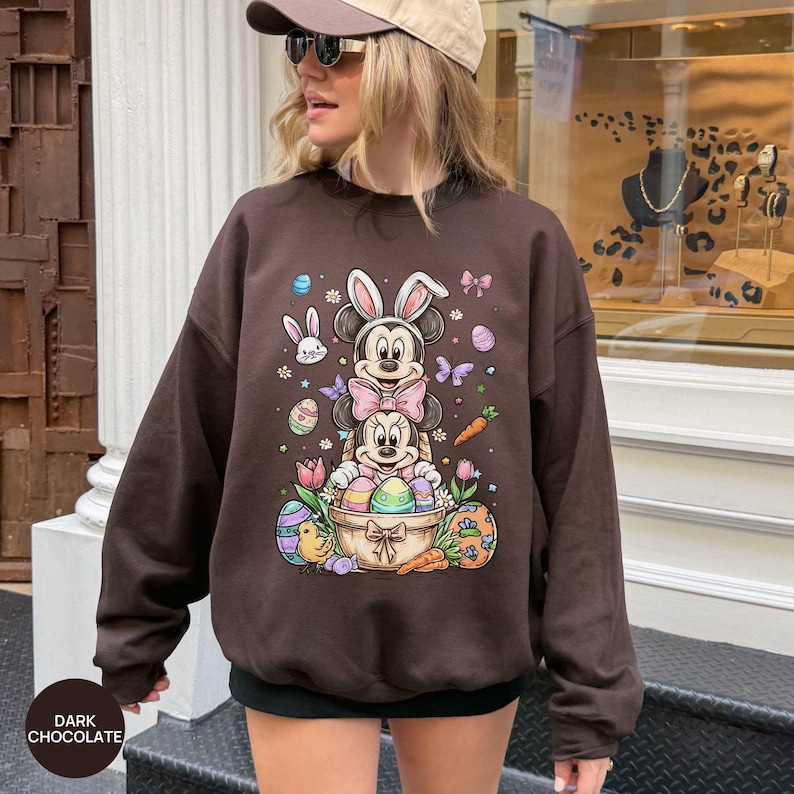 Puede incluir: Sudadera marr&oacute;n chocolate oscuro con un gr&aacute;fico colorido de Pascua. El gr&aacute;fico incluye los personajes de Mickey y Minnie Mouse con orejas de conejo, huevos de Pascua, zanahorias y mariposas. La sudadera tiene cuello redondo y mangas largas.