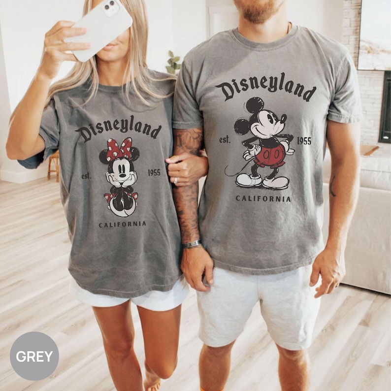 Puede incluir: Dos camisetas grises con el logotipo de Disneyland y gr&aacute;ficos de Mickey y Minnie Mouse. Las camisetas tienen el texto "Disneyland est. 1955 California". Las camisetas son de manga corta y tienen un aspecto vintage.