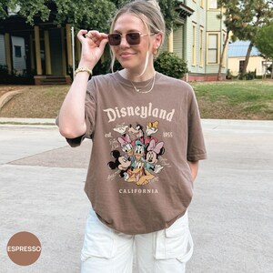 Puede incluir: Camiseta color espresso con el logo de Disneyland y Mickey Mouse, Minnie Mouse, Donald, Daisy, Goofy y Pluto. La camiseta tiene el texto "Disneyland 1955 California". La modelo lleva pantalones cargo blancos.
