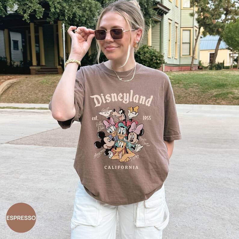 Puede incluir: Camiseta color espresso con el logo de Disneyland y Mickey Mouse, Minnie Mouse, Donald, Daisy, Goofy y Pluto. La camiseta tiene el texto "Disneyland 1955 California". La modelo lleva pantalones cargo blancos.