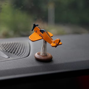 May include: A small, orange and blue airplane figurine with a propeller. The airplane is mounted on a silver spring and a wooden base. The figurine is displayed on a dark dashboard, with a blurred green background.