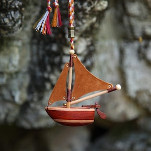 Könnte beinhalten: Ein Miniatur-Holzsegelschiff mit braunem Segel und rotem und braunem Rumpf, aufgehängt an einer bunten geflochtenen Schnur. Das Boot hat ein kleines Ruder und ist mit detaillierter Präzision gefertigt. Der Hintergrund ist eine verschwommene natürliche Umgebung.