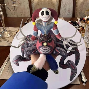 Puede incluir: Un peluche con una figura de bufón con una cara sonriente, atuendo rojo y azul, y un cuerpo de araña. El juguete se sostiene sobre un plato blanco, con una manga azul visible en primer plano.