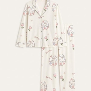 Puede incluir: Conjunto de pijama de dos piezas en color marfil con tem&aacute;tica de Pascua. El conjunto presenta una camisa abotonada de manga larga y pantalones a juego, ambos adornados con un patr&oacute;n repetido de conejitos dentro de huevos de Pascua, flores rosas y la palabra "Bunny".