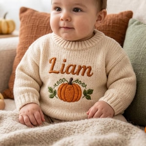 Könnte beinhalten: Ein cremefarbener, langärmeliger Strickpullover mit dem in brauner Schreibschrift gestickten Namen Liam über einer detaillierten orangefarbenen Kürbisgrafik mit grünen Blättern und Ranken, ausgestattet mit einem gerippten Rundhalsausschnitt.