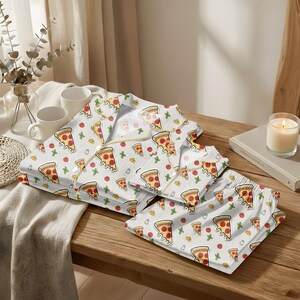 May include: White pajama set with a pizza slice pattern. The set includes a button-down shirt and shorts, both adorned with repeating illustrations of pizza slices, tomatoes, and green herbs. The pajamas are laid out on a wooden surface.