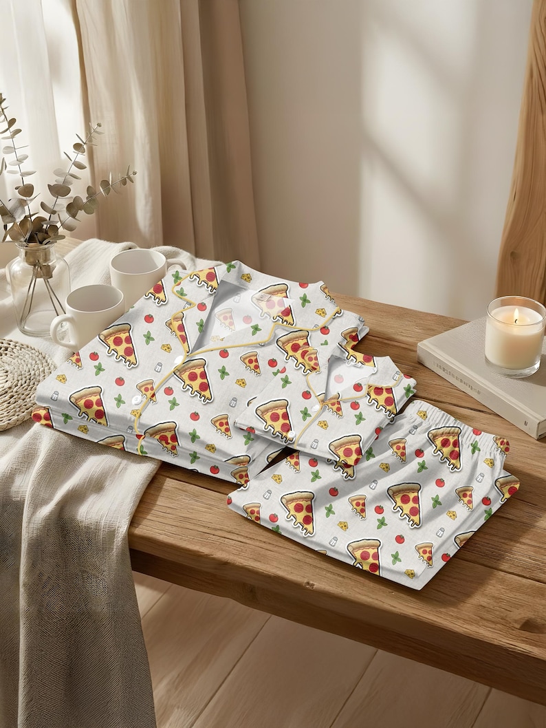 May include: White pajama set with a pizza slice pattern. The set includes a button-down shirt and shorts, both adorned with repeating illustrations of pizza slices, tomatoes, and green herbs. The pajamas are laid out on a wooden surface.