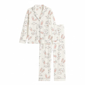 以下が含まれることがあります： ピンクの耳とアクセントが付いたウサギの顔の繰り返し模様が特徴の白いパジャマセット。 セットには、襟と長袖のボタンダウンのトップスと、お揃いのパンツが含まれています。 パジャマはグレーのトリムが付いています。