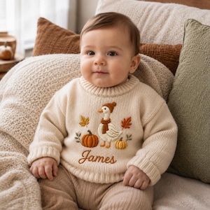 Könnte beinhalten: Ein cremefarbener Langarm-Strickpullover mit einer Stickerei, die eine Gans mit brauner Mütze und Schal zeigt, umgeben von orangefarbenen Kürbissen und Herbstblättern. Der Name James ist darunter in brauner Schreibschrift eingestickt.
