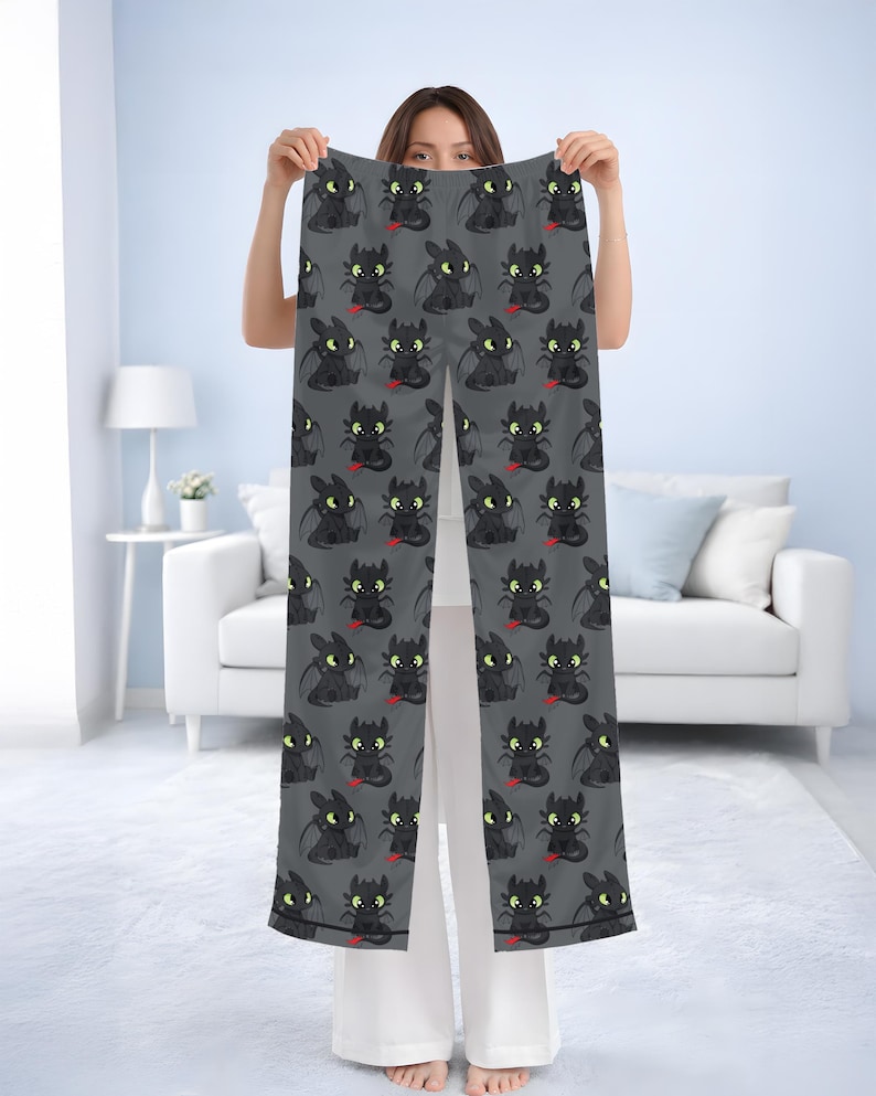 May include: Gray pajama pants with a repeating pattern of a cartoon dragon character. The dragon is black with yellow eyes and a red tongue. The pants are held up in front of a person.