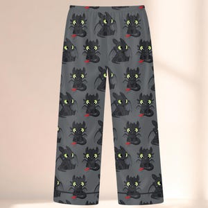 May include: Gray pajama pants featuring a repeating pattern of a cartoon dragon character. The dragons are black with green eyes and red accents. The pants have a relaxed fit and a soft appearance.