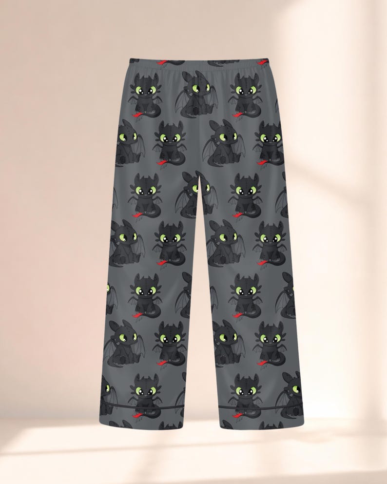 May include: Gray pajama pants featuring a repeating pattern of a cartoon dragon character. The dragons are black with green eyes and red accents. The pants have a relaxed fit and a soft appearance.