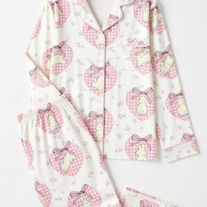 May include: White pajama set with a pink heart and bunny print. The set includes a button-down top with a chest pocket and matching pants. The word "PAJAMA" is written in pink letters at the top.