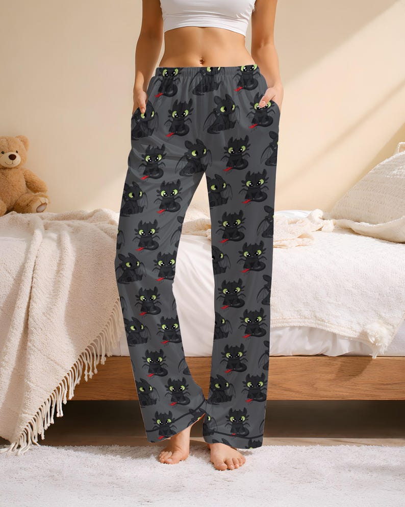 May include: Gray pajama pants with a repeating pattern of black cartoon dragons with red tongues and yellow eyes. The pants have a relaxed fit with pockets and a drawstring waist. The pajama pants are made of a soft, comfortable material.