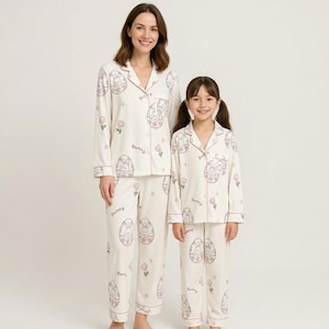 Puede incluir: Pijamas marfil a juego con un estampado de conejito y huevo, usados por una mujer y una ni&ntilde;a. Los pijamas tienen una parte superior abotonada y pantalones, con ribetes en rosa suave. La palabra "Bunny" est&aacute; impresa en los pantalones.