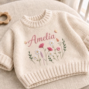 Può includere: Un maglione in maglia color crema con il nome Amelia ricamato in corsivo rosa, circondato da un motivo floreale con margherite bianche, fiori rosa, steli verdi e due piccole farfalle gialle sulla parte del petto.