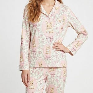 Puede incluir: Un conjunto de pijama rosa claro con un patr&oacute;n repetido de conejitos dentro de &oacute;valos rosas con lazos, sobre un fondo floral. La parte superior de manga larga tiene cuello y bolsillo en el pecho. El pantal&oacute;n de pijama tiene un dise&ntilde;o a juego.