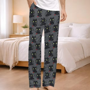 May include: Gray pajama pants featuring a repeating pattern of a black dragon with green eyes and a red tongue. The pajama pants are made of a soft material and are designed for comfort. The pants are shown on a person standing in a bedroom.