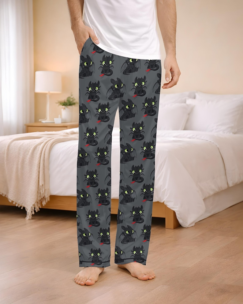 May include: Gray pajama pants featuring a repeating pattern of a black dragon with green eyes and a red tongue. The pajama pants are made of a soft material and are designed for comfort. The pants are shown on a person standing in a bedroom.
