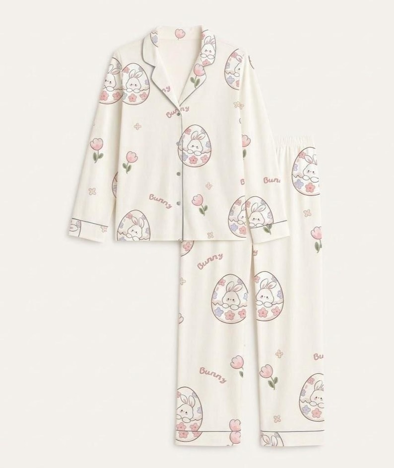 Puede incluir: Conjunto de pijama de dos piezas en color marfil con tem&aacute;tica de Pascua. El conjunto presenta una camisa abotonada de manga larga y pantalones a juego, ambos adornados con un patr&oacute;n repetido de conejitos dentro de huevos de Pascua, flores rosas y la palabra "Bunny".